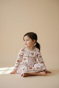 A young girl with dark hair in a ponytail sits cross-legged on a warm beige studio floor, wearing a long-sleeve cream dress with a brown patchwork pattern and small pumpkin and bows motifs.