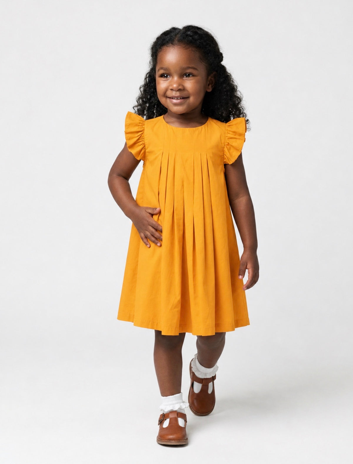 A toddler Black girl smiling while wearing a marigold-colored, pleated flutter-sleeve dress, white ruffle socks, and brown shoes