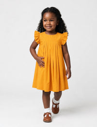 A toddler Black girl smiling while wearing a marigold-colored, pleated flutter-sleeve dress, white ruffle socks, and brown shoes